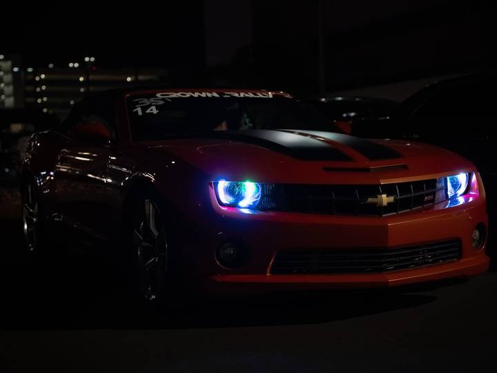 An orange Chevrolet Camaro at night with glowing blue headlight rings