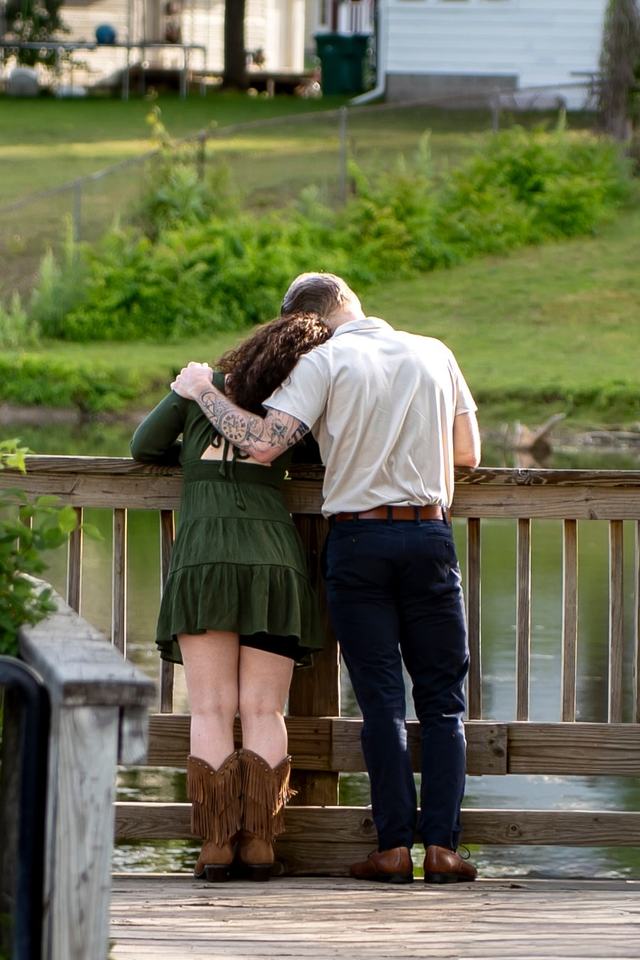 A couple leans on a wooden dock railing, arms around each other, looking over a pond