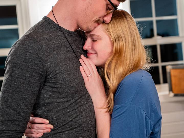 A couple embraces by a window, her eyes closed and his lips on her forehead