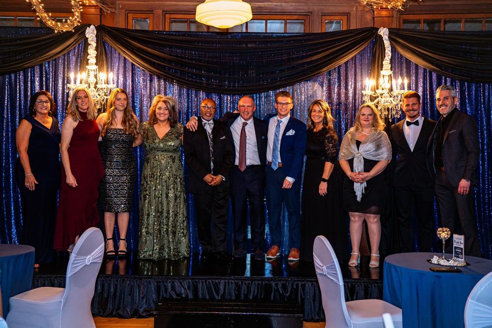 Guests in evening wear stand together on a stage under chandeliers and blue drapes
