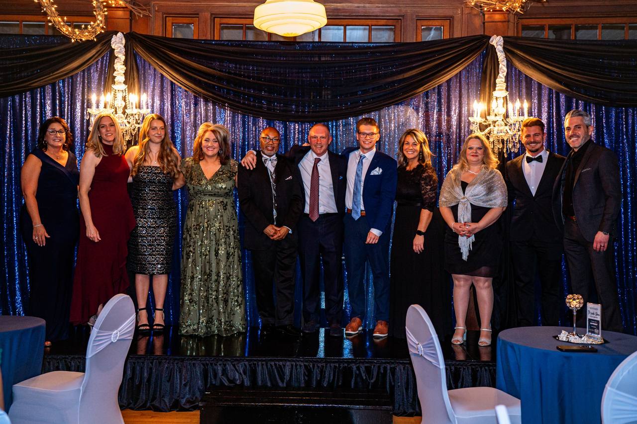 Guests in evening wear stand together on a stage under chandeliers and blue drapes