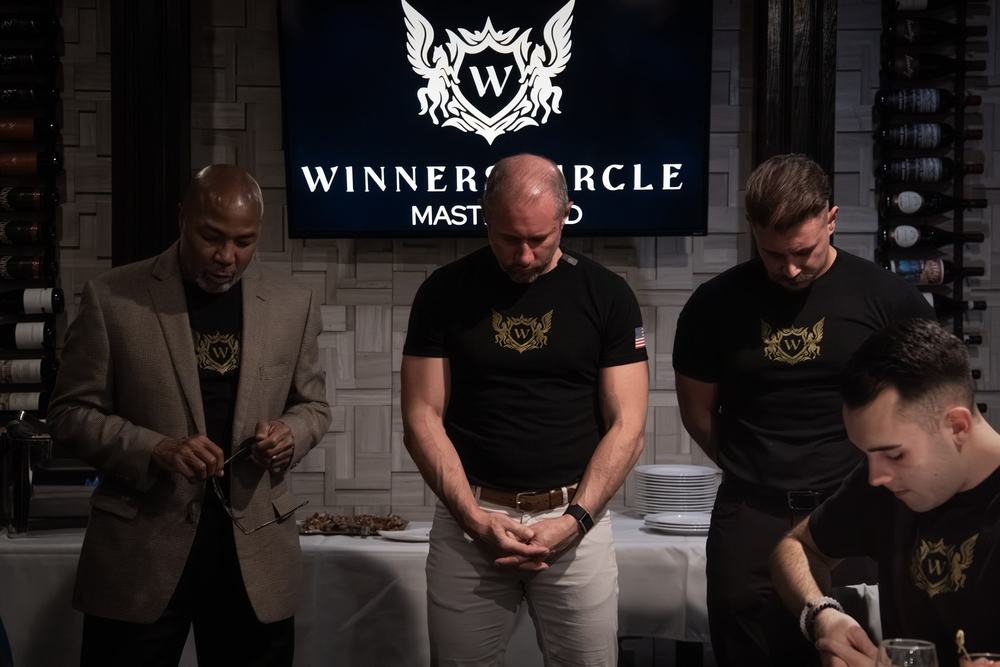 Men in black shirts stand with heads bowed beneath a Winners Circle Mastermind sign