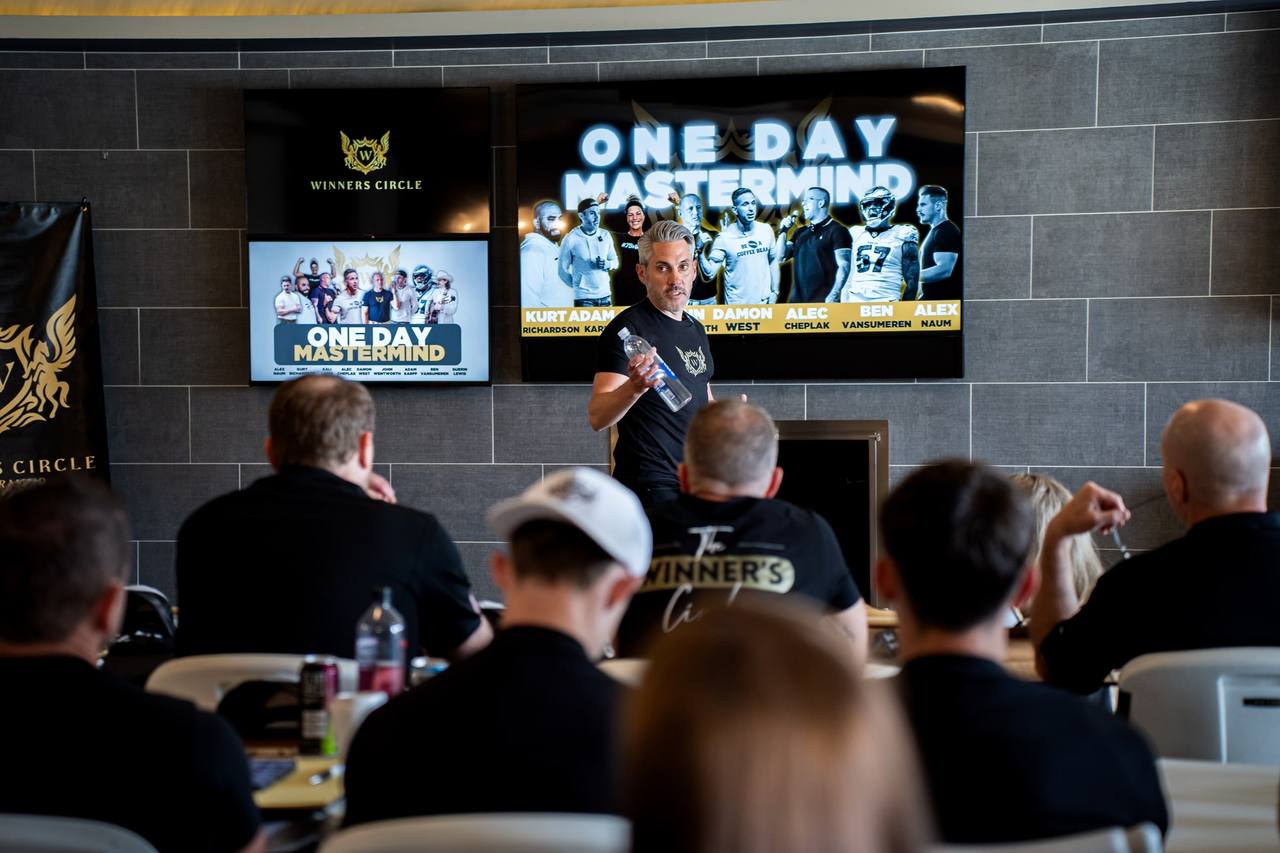 A speaker addresses a seated room in front of screens reading One Day Mastermind