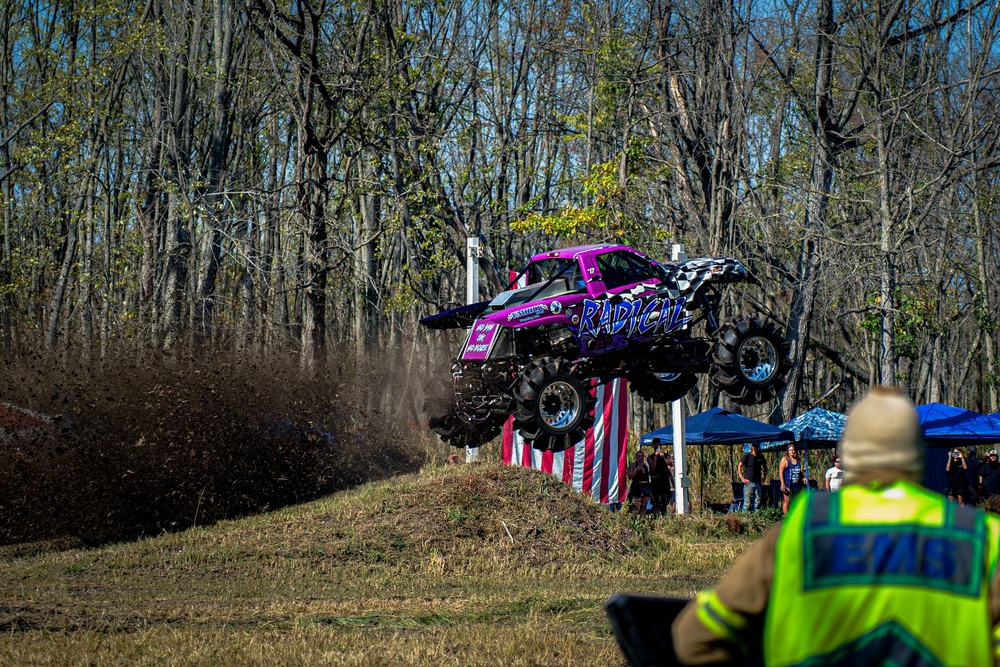 A purple monster truck in mid-air over a dirt jump as a safety worker watches