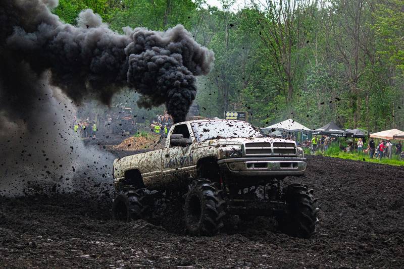 A mud-covered pickup on huge tires churns through a mud pit, black smoke pouring from its stack