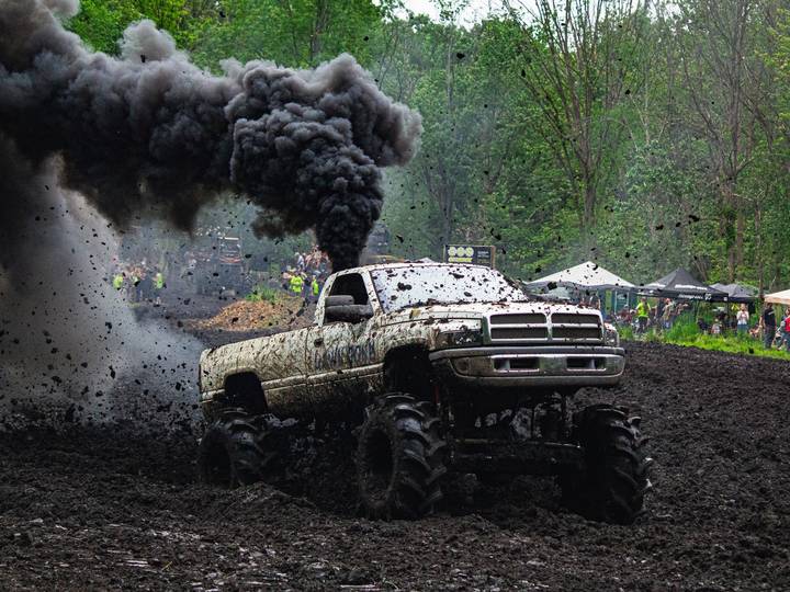A mud-covered pickup on huge tires churns through a mud pit, black smoke pouring from its stack