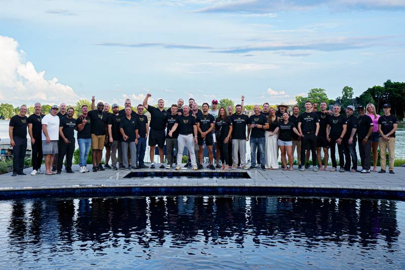 A large group in matching black shirts poses on a pool deck at sunset, mirrored in the water