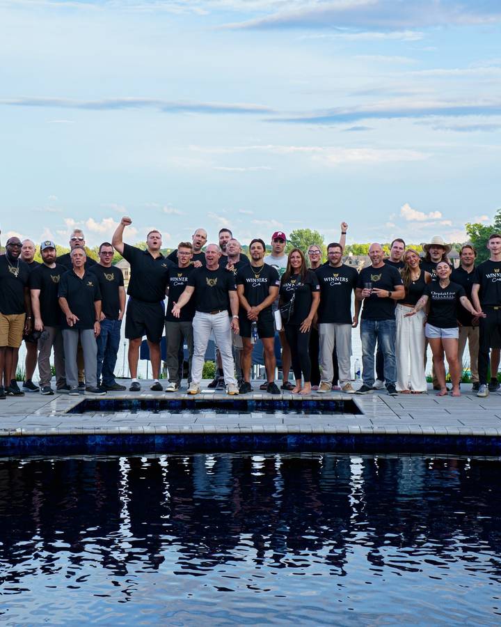 A large group in matching black shirts poses on a pool deck at sunset, mirrored in the water