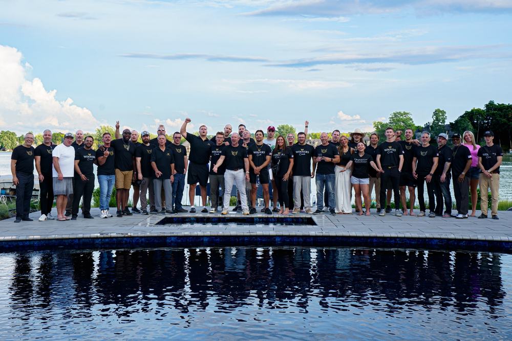 A large group in matching black shirts poses on a pool deck at sunset, mirrored in the water