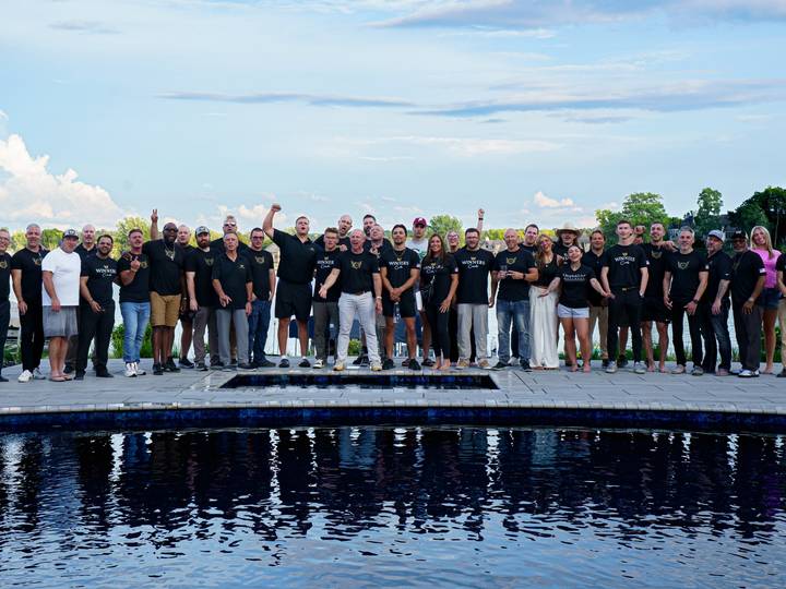 A large group in matching black shirts poses on a pool deck at sunset, mirrored in the water