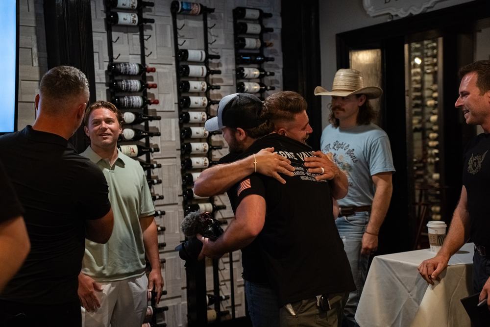 Guests greet each other with hugs and handshakes in a room lined with wine racks