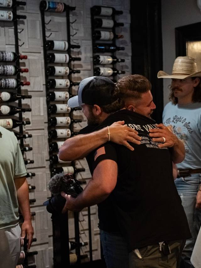 Guests greet each other with hugs and handshakes in a room lined with wine racks