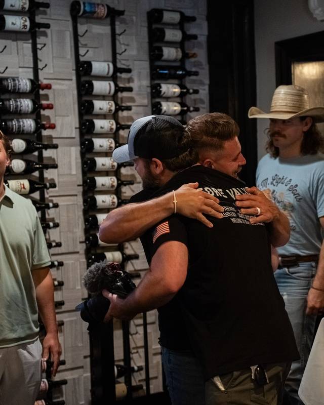 Guests greet each other with hugs and handshakes in a room lined with wine racks