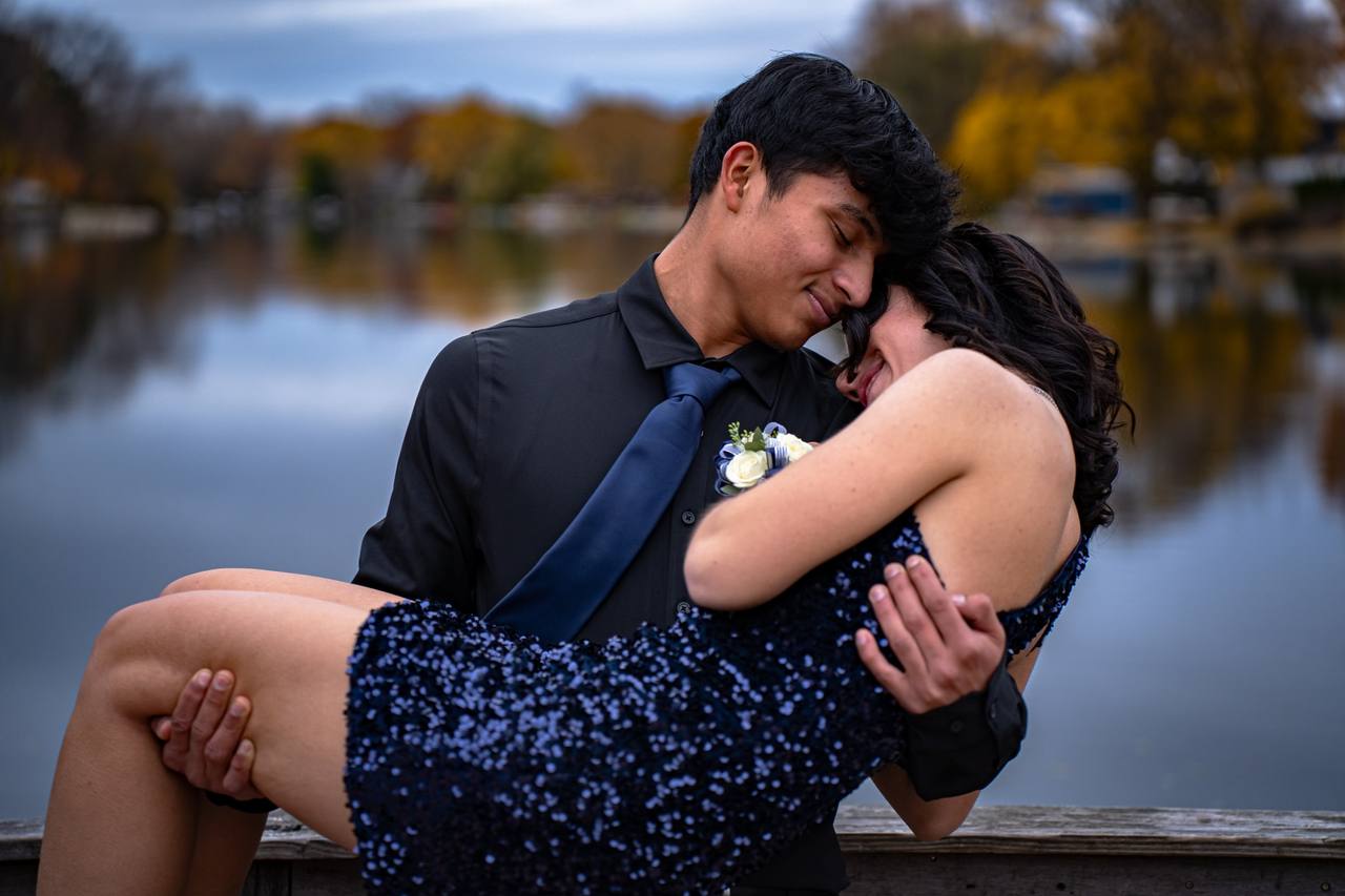 A young man in a dark suit holds a young woman in a sequined dress in front of a lake