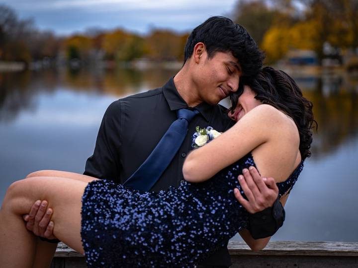 A young man in a dark suit holds a young woman in a sequined dress in front of a lake