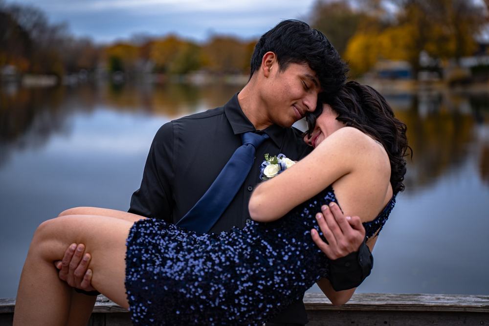 A young man in a dark suit holds a young woman in a sequined dress in front of a lake