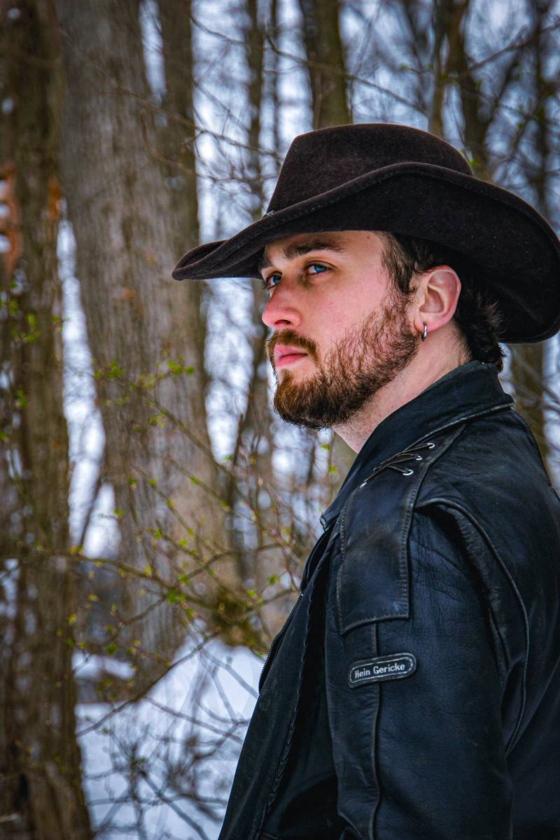 A bearded man in a black cowboy hat and leather jacket looks off to the side among bare trees