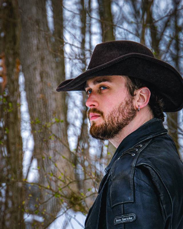 A bearded man in a black cowboy hat and leather jacket looks off to the side among bare trees