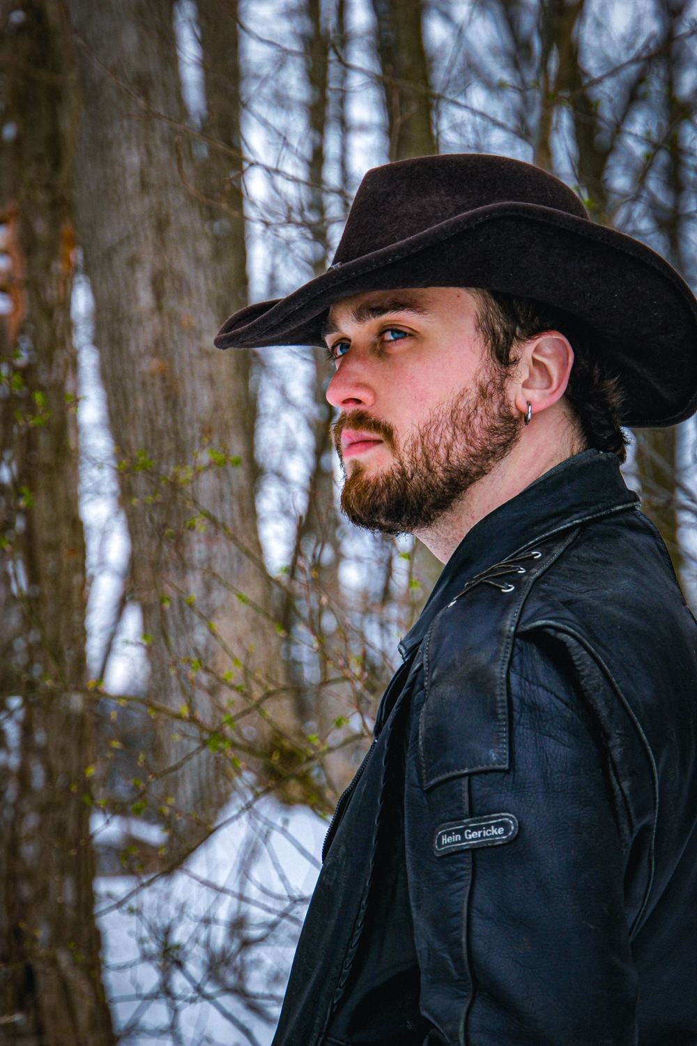 A bearded man in a black cowboy hat and leather jacket looks off to the side among bare trees