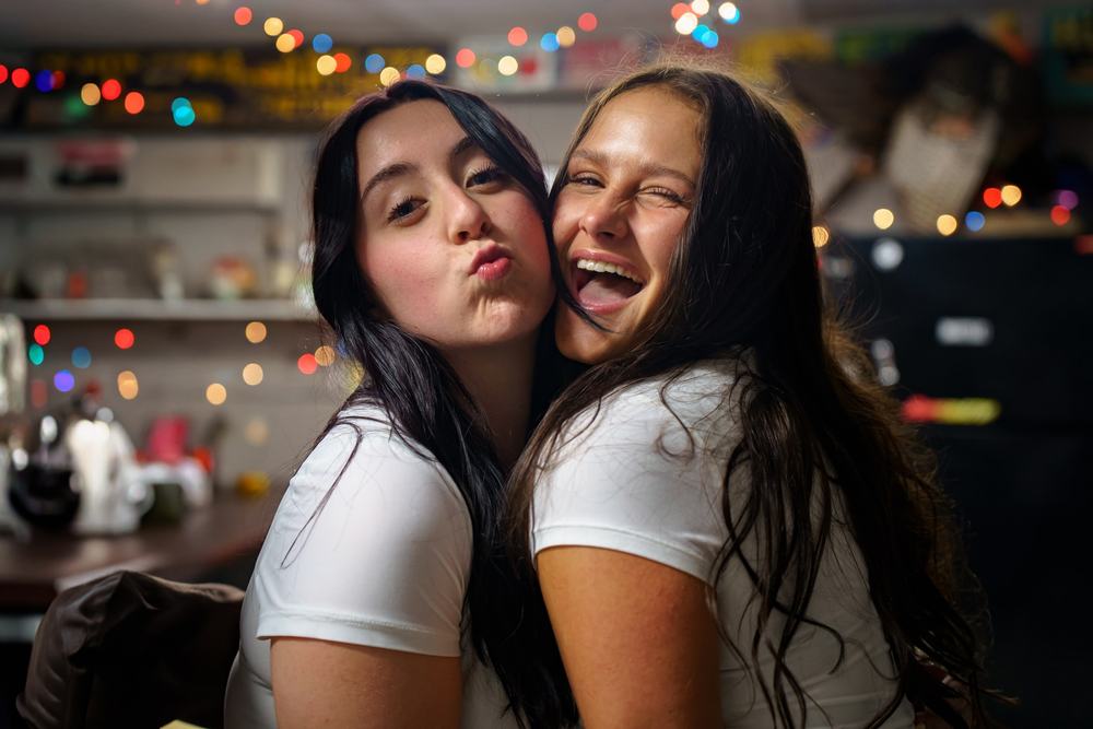 Two friends squeeze together and pull faces in front of colorful string lights