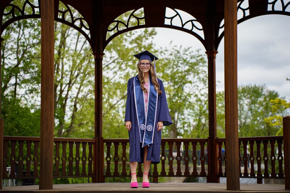 A graduate in cap, gown and stoles stands in the center of a wooden gazebo