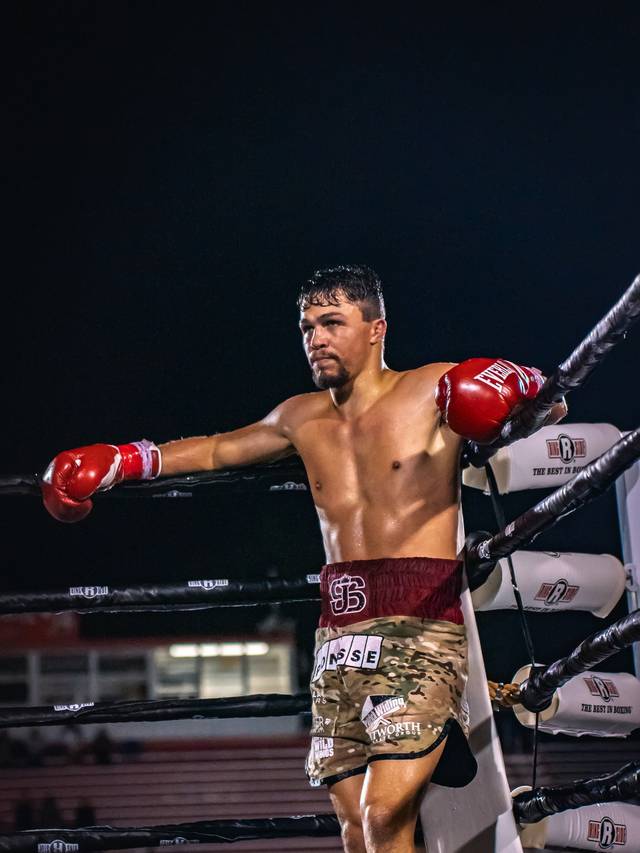 A boxer in red gloves leans on the ropes in his corner, a crowd behind him