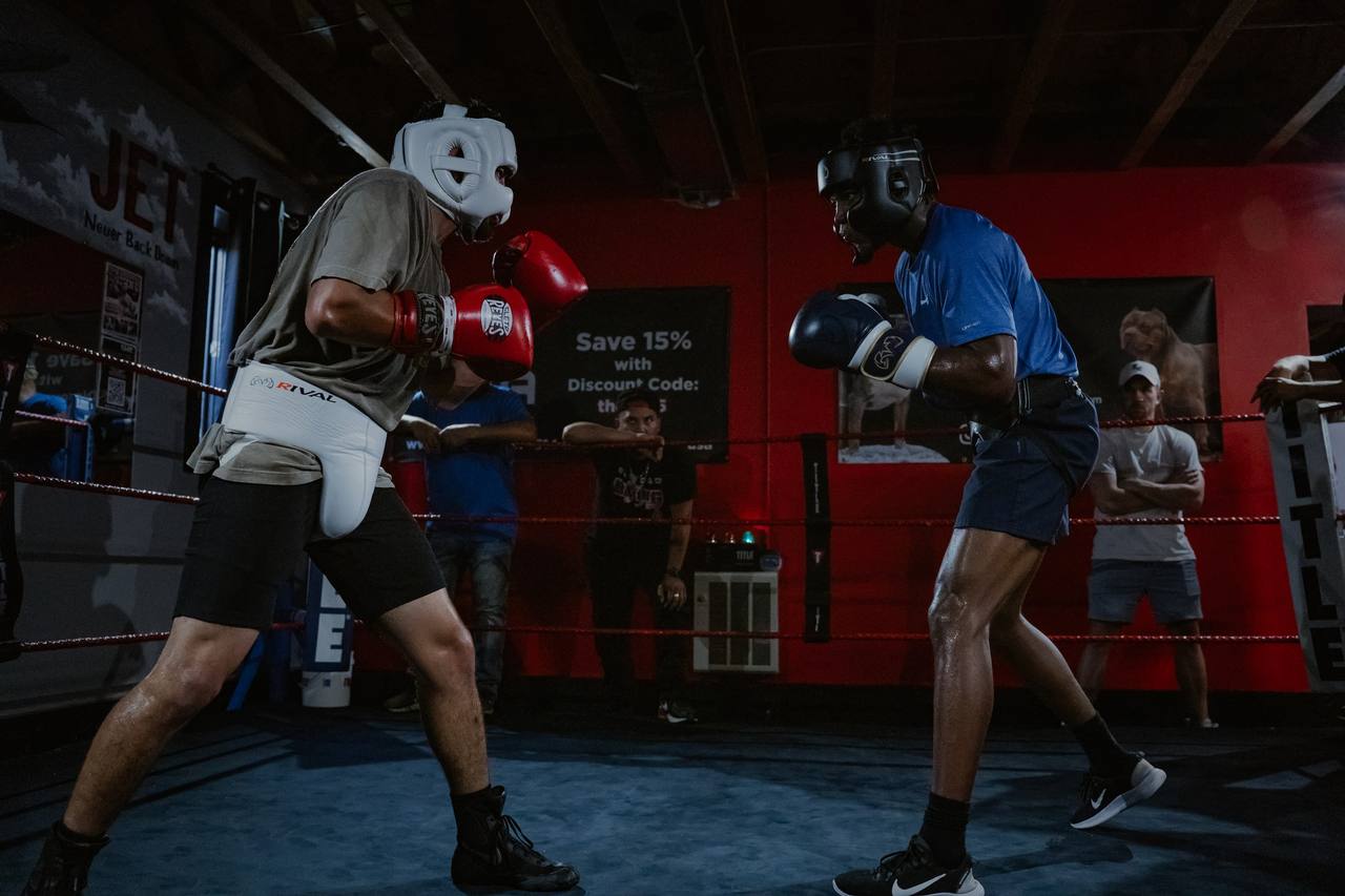 Two fighters in headgear spar in a gym ring with a red wall behind them