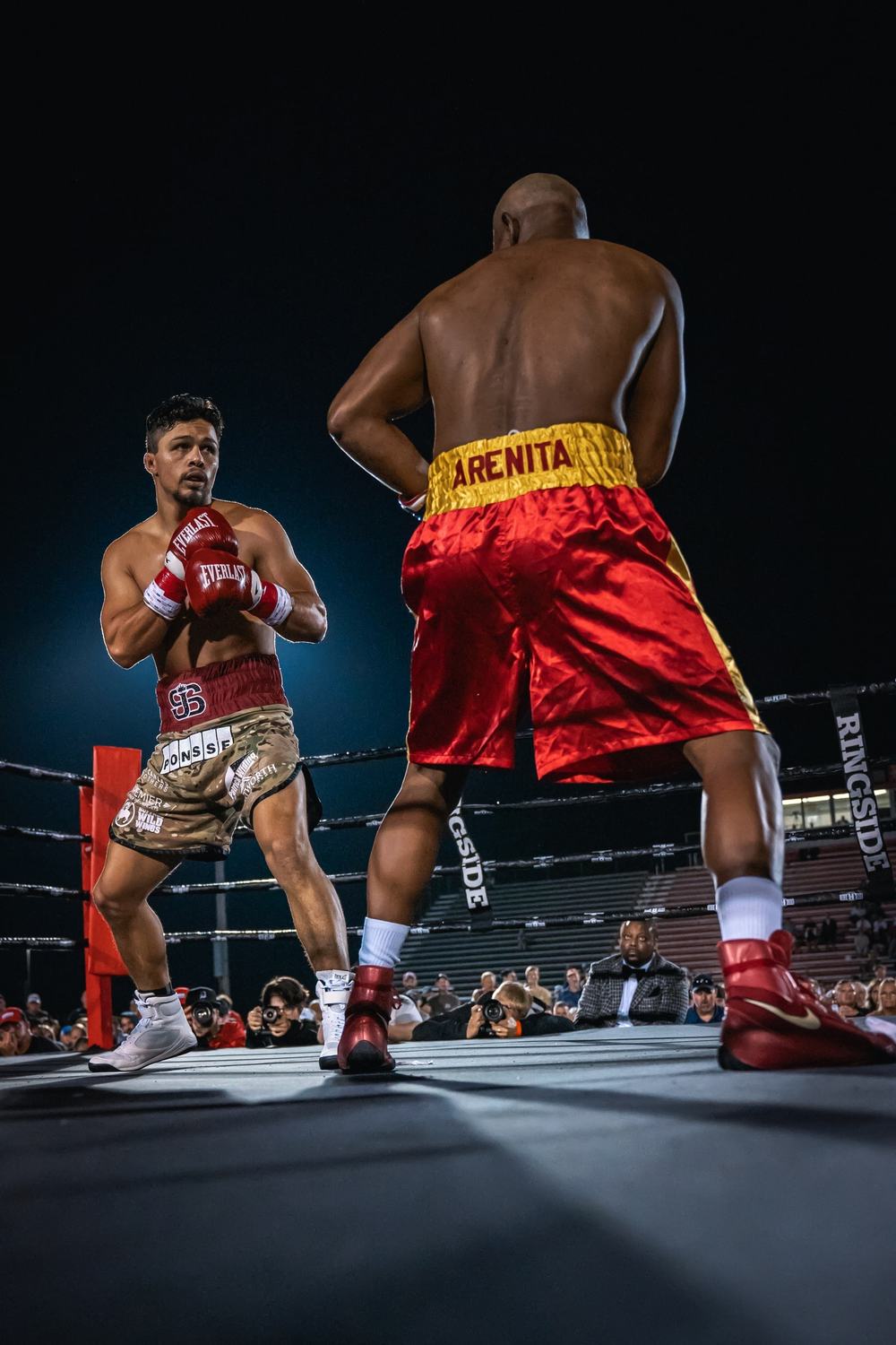 Two boxers square off in an outdoor ring at night, seen from ring level