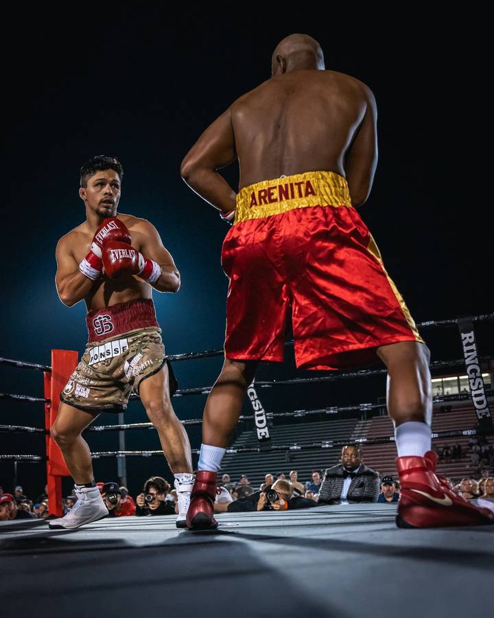 Two boxers square off in an outdoor ring at night, seen from ring level