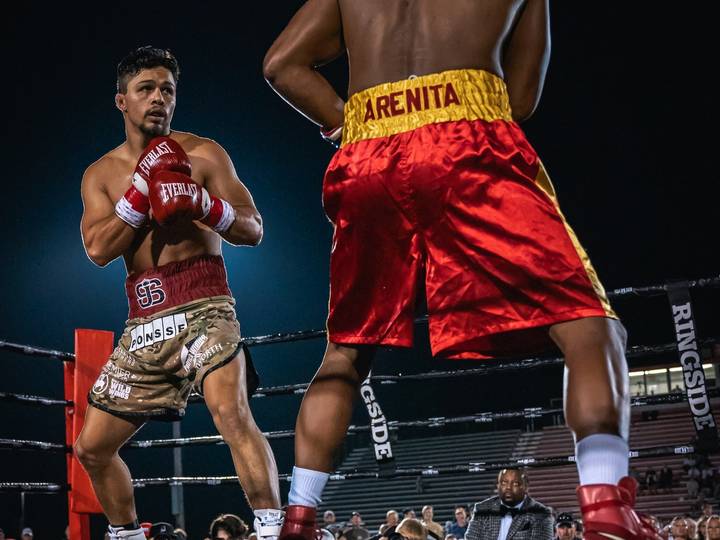 Two boxers square off in an outdoor ring at night, seen from ring level